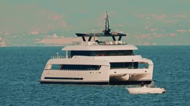 Arka planda kıyı kenti olan berrak mavi sulara demirlemiş modern beyaz bir katamaran yatı.