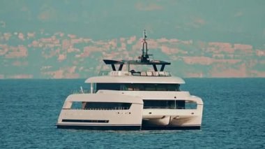 Arka planda kıyı kenti olan berrak mavi sulara demirlemiş modern beyaz bir katamaran yatı.