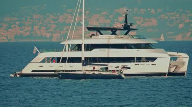 Antibes, France - November 10, 2025: A luxury yacht anchored offshore with a sailboat gliding past in the foreground, set against a scenic coastal skyline