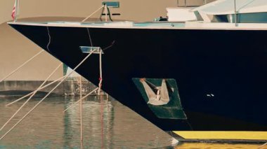 Bir yatın pruvasına yakın durgun marina suyunda parıldayan bir çapa ve sarı gövde yansıması görünüyor.