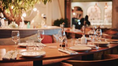 Sıcak ışıklandırma, ahşap mobilyalar, şarap kadehleri ve rafine masa düzeniyle samimi bir restoranın sinematik çekimi.