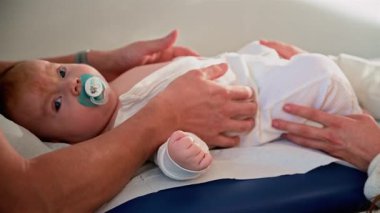 Bir bebeğin tıbbi muayene masasında sakince yatarken, elinde bir emzik tutarken bir yetişkinin nazikçe onları desteklemesi.