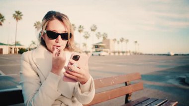 Güneş gözlüğü takan zarif bir kadın gün batımında bankta otururken ruj sürüyor.