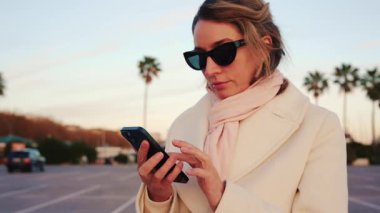 Beyaz önlüklü, eşarplı ve güneş gözlüklü zarif bir kadın telefonunu kontrol ediyor. Palmiye ağaçlarıyla kaplı güneşli bir alanda.