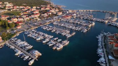Yat ve yelkenli teknelerle dolu, kırmızı çatılı evleri olan kıyı kasabasıyla çevrili büyük bir marinanın insansız hava aracı görüntüsü. Tribunj, Hırvatistan