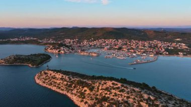 Aerial drone view of rocky green islands separated by narrow channels of turquoise water, with a coastal town and marina visible in the background. Tribunj, Croatia