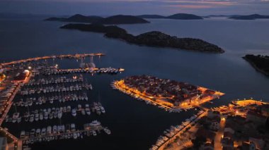 Tribunj Adası 'nın küçük bir kasabasını çevreleyen ve sıcak sokak lambalarıyla aydınlatılan teknelerle dolu parlayan bir marinanın insansız hava aracı görüntüsü.