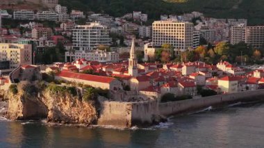 Budva Eski Kasabası 'nın hava aracı görüntüsü, ortaçağ taş binalarını, kırmızı çatılarını, kale duvarlarını ve kıyı şeridi boyunca bulunan kilise kulesini, modern şehir binalarını ve arka planında dağları gösteriyor. Karadağ