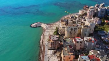 Durres sahil şeridinin hava aracı görüntüsü uzun apartmanlar, turkuaz deniz ve suya uzanan eşsiz şekilli liman platformunu gösteriyor.