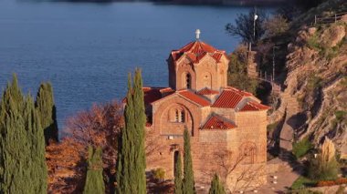 Aerial drone view of the iconic St. John the Theologian Church perched above Lake Ohrid, framed by cypress trees and autumn colors. North Macedonia