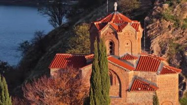 Aerial drone view of the iconic St. John the Theologian Church perched above Lake Ohrid, framed by cypress trees and autumn colors. North Macedonia
