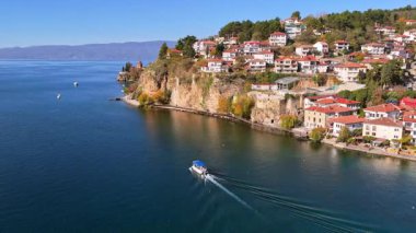 Göl teknesinin insansız hava aracı görüntüsü Kaneo kayalıklarına doğru ilerliyor. Arka planda St. John Kilisesi ve deniz manzaralı tepedeki büyüleyici evler görülüyor.