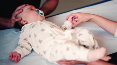Bir yetişkinin bebeğin elini nazikçe tutarken diğer bir bakıcı da sağlık kontrolü sırasında bebeğin başını desteklerken.