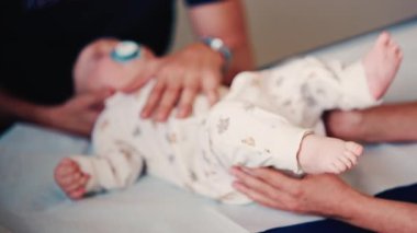 Bir yetişkinin bebeğin elini nazikçe tutarken diğer bir bakıcı da sağlık kontrolü sırasında bebeğin başını desteklerken.