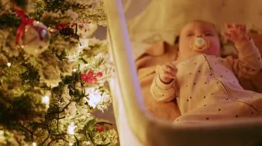 Sıcak ve samimi bir Noel sahnesinde beşikte huzur içinde uzanan bir bebek hafifçe aydınlatılmış bir Noel ağacının yanında