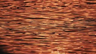 Turuncu ve altın gün batımının yansımaları yumuşak su dalgaları arasında hareket eder.