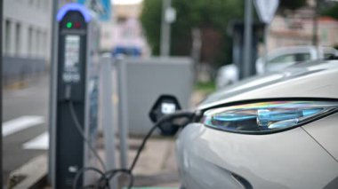 Kentsel bir çevrede beyaz, elektrikli bir araç halka açık bir şarj istasyonuna park edilmiş.