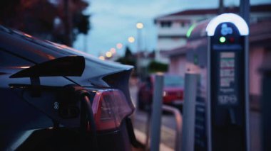 Akşam saatlerinde halka açık bir şarj istasyonuna takılan siyah, elektrikli bir araba.