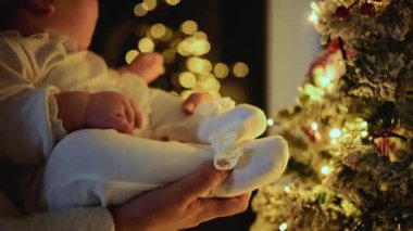 Süslü bir Noel ağacına yakın tutulan bebek, aydınlatılmış süs ve ışıklara doğru uzanıyor.