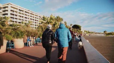 Cannes, Fransa - 23 Aralık 2025: Güneşli bir günde, arka planda palmiye ağaçları, banklar ve Akdeniz ile La Croisette boyunca yürüyen yayalar