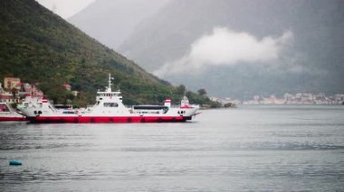 Güvertede arabalı feribot, arka planda dağ ve kıyı kasabası olan körfezi geçiyor.