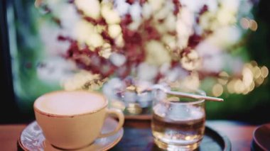 Kahve fincanı pencerenin yakınındaki bir masaya koyulmuş. Arka planda yumuşak bokeh ışıkları ve bulanık yapraklar var.