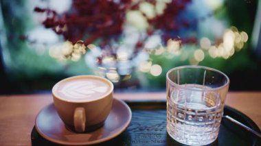 Kahve fincanı pencerenin yakınındaki bir masaya koyulmuş. Arka planda yumuşak bokeh ışıkları ve bulanık yapraklar var.