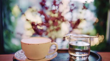 Kahve fincanı pencerenin yakınındaki bir masaya koyulmuş. Arka planda yumuşak bokeh ışıkları ve bulanık yapraklar var.