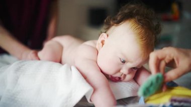 Bebek, yumuşak bir yastığın üzerinde karnının üzerinde yatarken, bakıcı tarafından desteklenen, hareket egzersizleri yapan küçük bir oyuncağa uzanıyor.