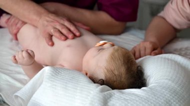 Bebek sırt üstü yatarken bir yetişkin onlara masaj yapıyor ve anne nazikçe bebeğin elini tutuyor.
