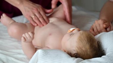 Bebek sırt üstü yatarken bir yetişkin onlara masaj yapıyor ve anne nazikçe bebeğin elini tutuyor.