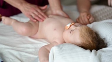 Bebek sırt üstü yatarken bir yetişkin onlara masaj yapıyor ve anne nazikçe bebeğin elini tutuyor.