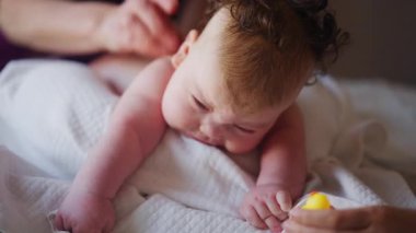 Masaj sırasında oyuncak ile battaniyeye uzanmış ağlayan bir bebeğin dikkatini dağıtmaya çalışan bir kadın.