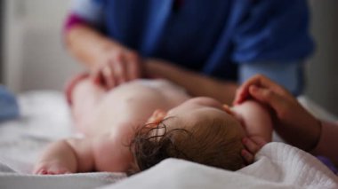 Bebek sırt üstü yatarken bir yetişkin onlara masaj yapıyor ve anne nazikçe bebeğin elini tutuyor.