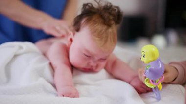 Masaj sırasında oyuncak ile battaniyeye uzanmış ağlayan bir bebeğin dikkatini dağıtmaya çalışan bir kadın.