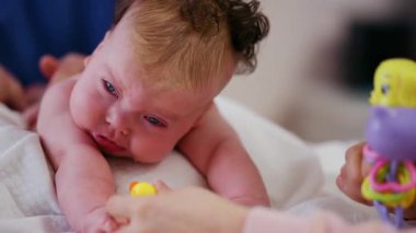 Masaj sırasında oyuncak ile battaniyeye uzanmış ağlayan bir bebeğin dikkatini dağıtmaya çalışan bir kadın.