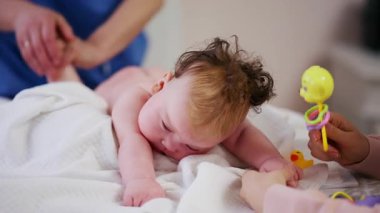 Masaj sırasında oyuncak ile battaniyeye uzanmış ağlayan bir bebeğin dikkatini dağıtmaya çalışan bir kadın.