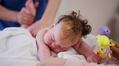 Masaj sırasında oyuncak ile battaniyeye uzanmış ağlayan bir bebeğin dikkatini dağıtmaya çalışan bir kadın.