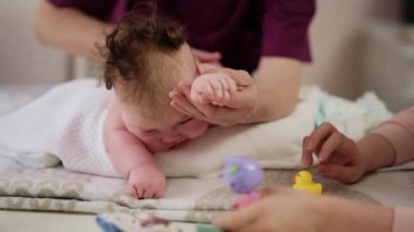 Masaj sırasında oyuncak ile battaniyeye uzanmış ağlayan bir bebeğin dikkatini dağıtmaya çalışan bir kadın.