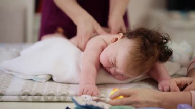 Masaj sırasında oyuncak ile battaniyeye uzanmış ağlayan bir bebeğin dikkatini dağıtmaya çalışan bir kadın.