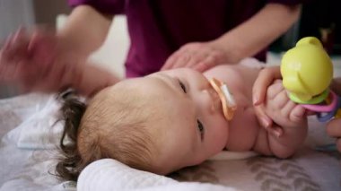 Masaj sırasında oyuncak ile battaniyeye uzanmış ağlayan bir bebeğin dikkatini dağıtmaya çalışan bir kadın.