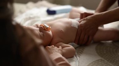 Bebek sırt üstü yatarken bir yetişkin onlara masaj yapıyor ve anne nazikçe bebeğin elini tutuyor.