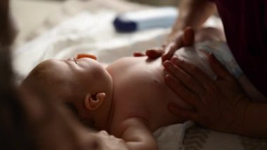 Bebek sırt üstü yatarken bir yetişkin onlara masaj yapıyor ve anne nazikçe bebeğin elini tutuyor.