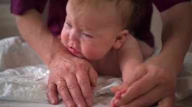 Yumuşak bir yüzeye yerleştirilmiş bebek, yetişkinler tarafından nazikçe desteklenen karın egzersizi yapıyor.