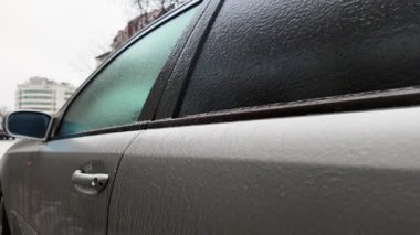 Buz ve buzla kaplı bir arabanın yan görüntüsü, kentsel arka planda desenli donmuş bir yüzey yaratıyor.