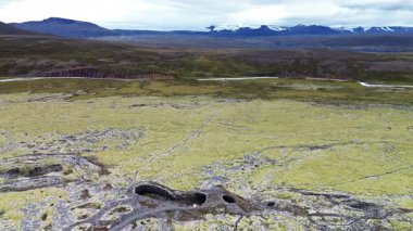 Hava aracı geniş bir yosun tabakasını volkanik mağaralar ve araziye yayılmış açıklıklarla kaplıyor. İzlanda dağlarının ufuklarında uzak dağlar ve buzullar görülebilir.