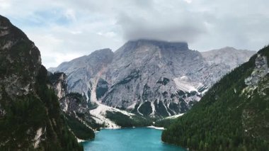 Sarp kayalık dağlar ve yoğun çam ormanlarıyla çevrili turkuaz bir alp gölü hava aracı görüntüsü. Bulutlar Dolomitlerin yüksek tepelerini kısmen kaplar