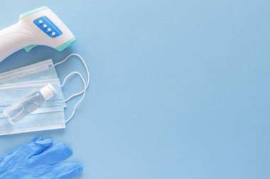Corona virüsü koruma konsepti düzdü. Temassız kızılötesi vücut termometresi, lateks eldivenler, tek kullanımlık maskeler ve metin alanı için mavi arkaplan üzerinde dezenfektan..