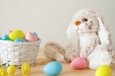 Paskalya oyuncak tavşanı, sepetli renkli yumurtalar ve ahşap masa üzerinde beyaz duvar zeminli küçük çiçekler ve metin için kopyalama alanı.