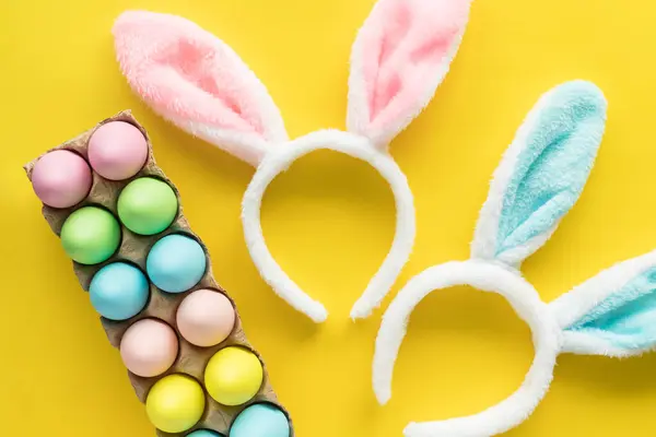 Colored Easter eggs and bunny ears headbands for children. Easter holiday flat lay on yellow background. Egg hunting concept
